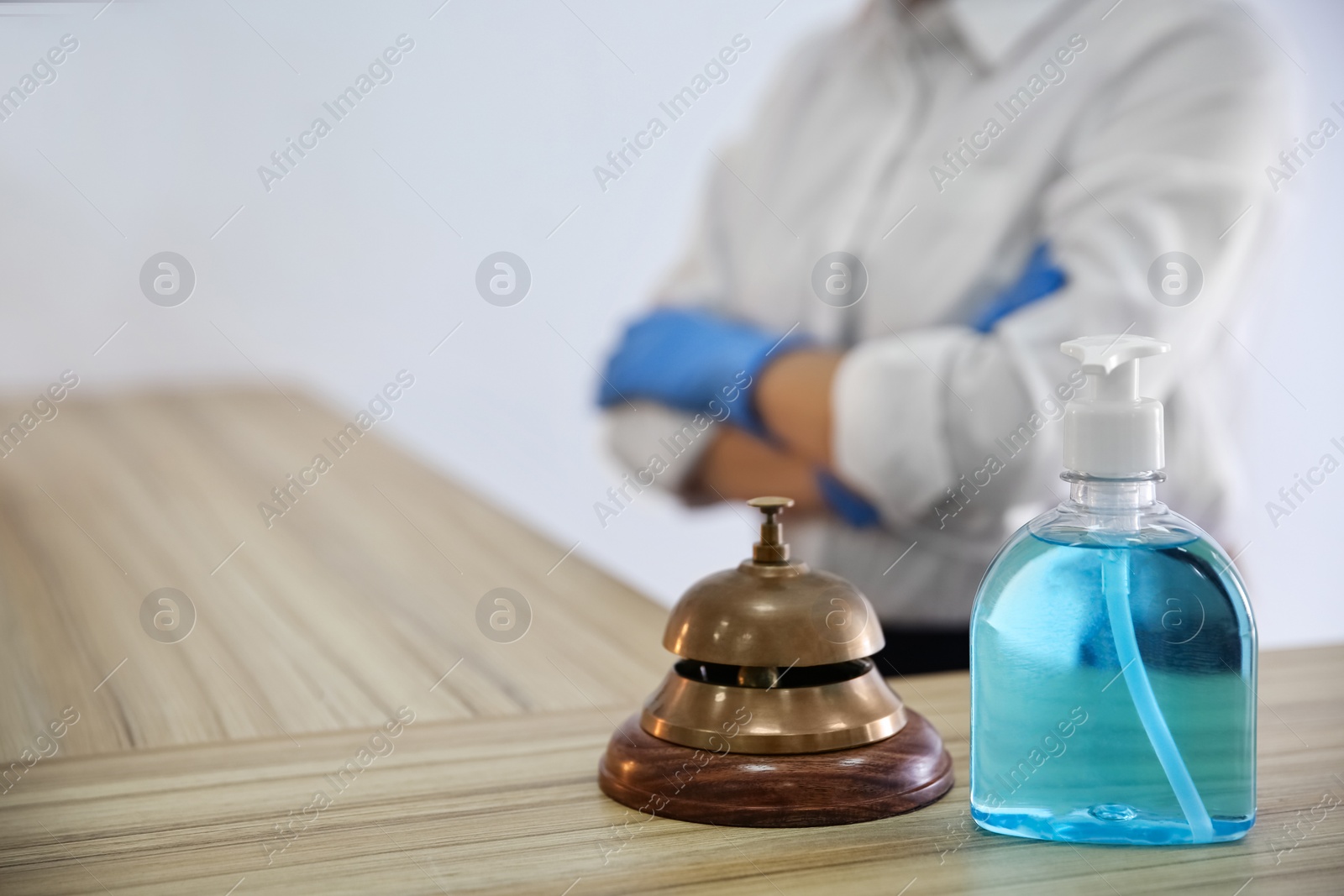 Photo of Receptionist at countertop in hotel, focus on dispenser bottle with antiseptic gel and service bell. Space for text