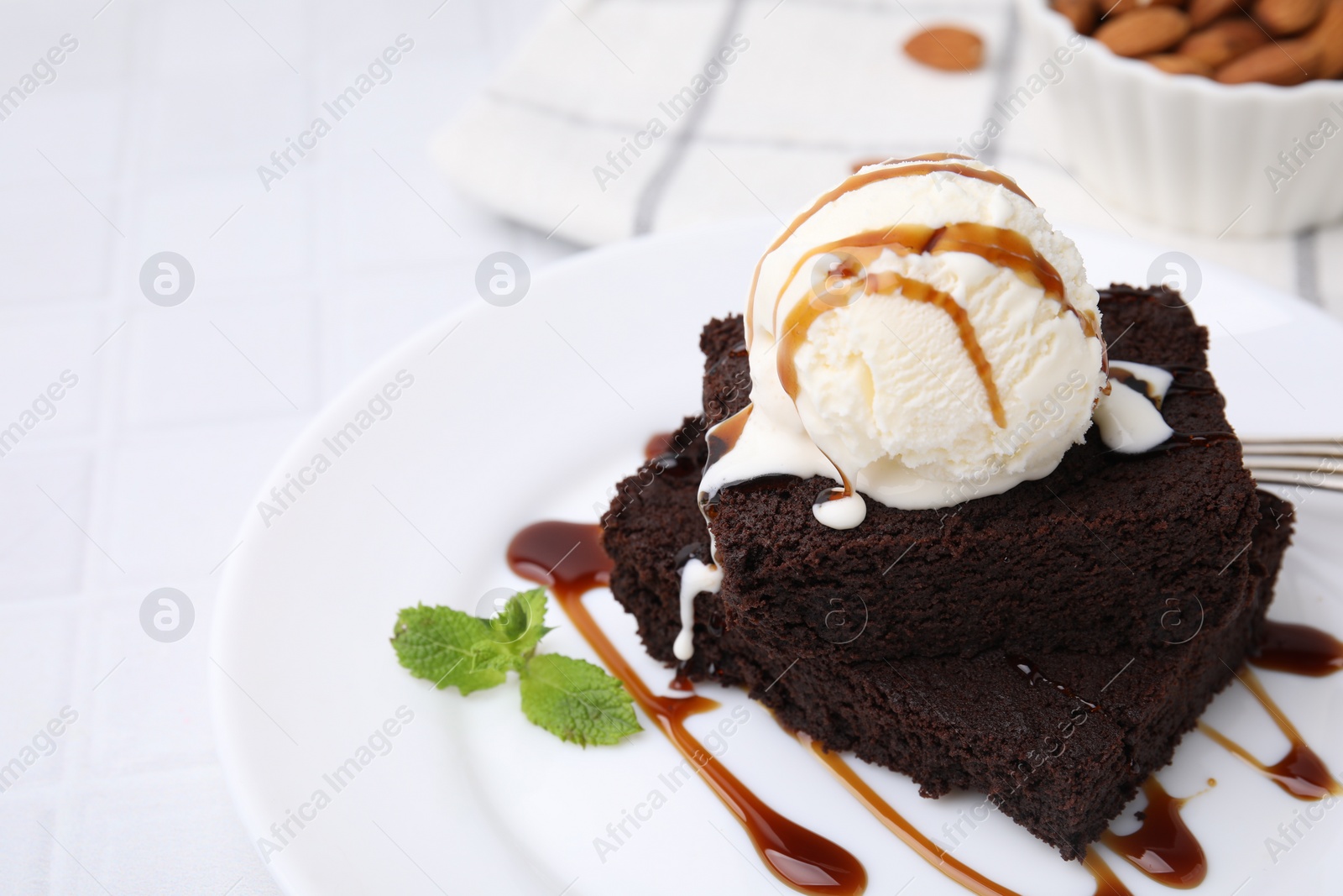 Photo of Tasty brownies served with ice cream and caramel sauce on white tiled table, closeup. Space for text