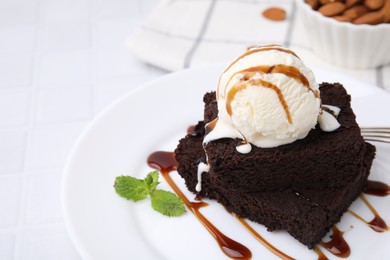 Photo of Tasty brownies served with ice cream and caramel sauce on white tiled table, closeup. Space for text