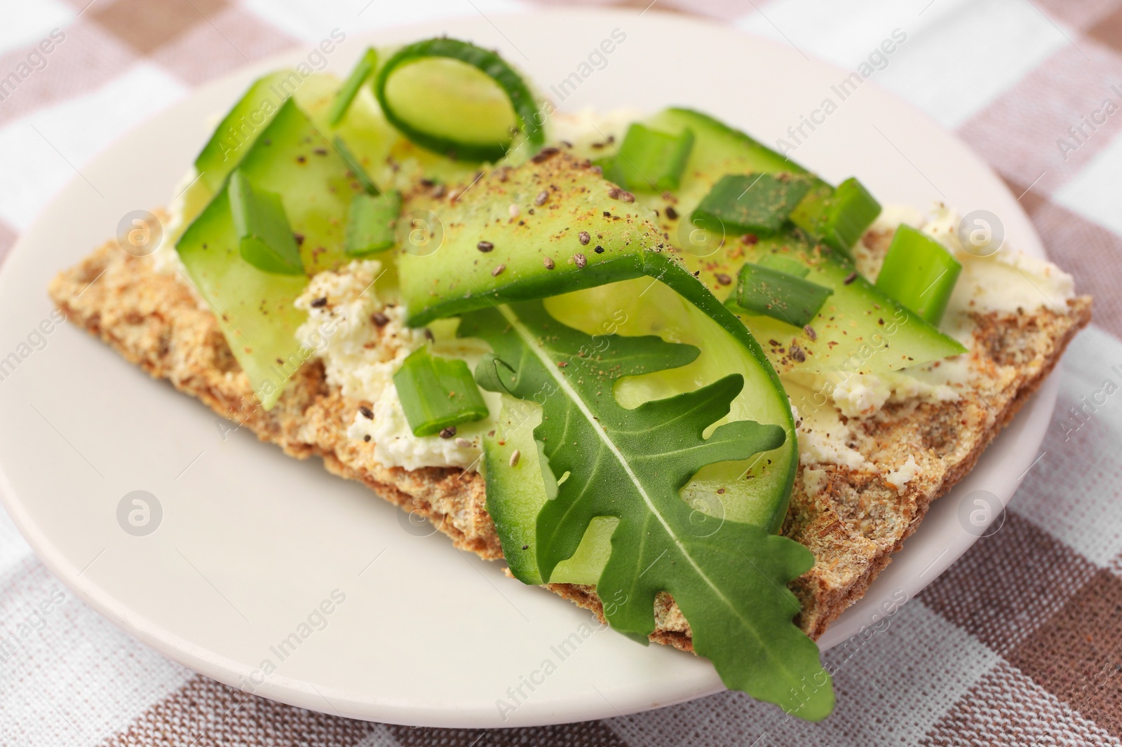 Photo of Fresh crunchy crispbread with cream cheese, cucumber, green onion and arugula on checkered tablecloth, closeup