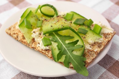 Photo of Fresh crunchy crispbread with cream cheese, cucumber, green onion and arugula on checkered tablecloth, closeup