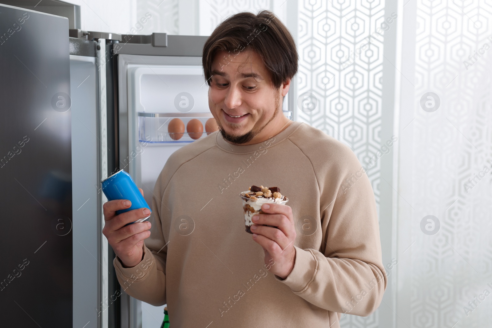 Photo of Overweight man holding dessert and tin can with beverage near open refrigerator in kitchen