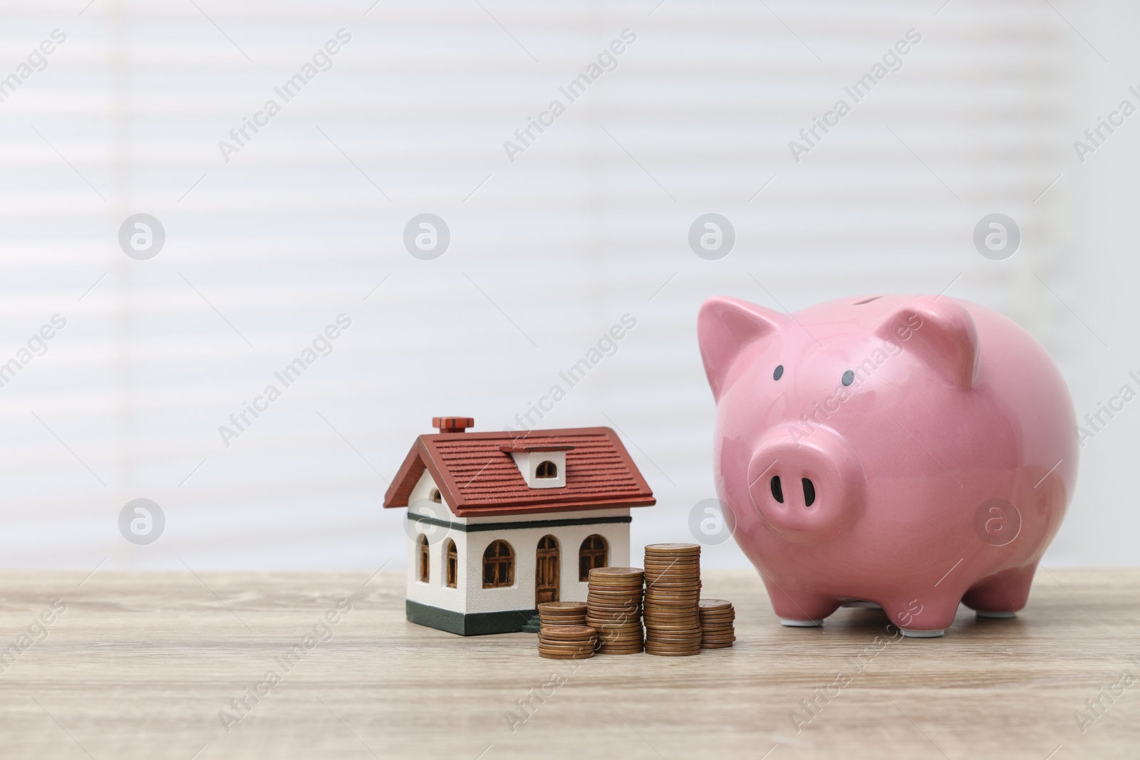 Photo of House model, piggy bank and stacked coins on light wooden table, space for text