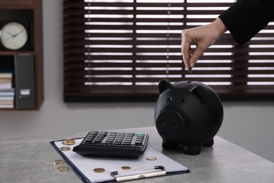 Woman putting money into piggy bank on grey table in office, closeup