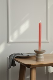 Holder with burning candle on wooden table near white wall indoors