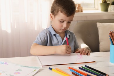 Photo of Cute little boy drawing with pencil at white wooden table in room. Child`s art
