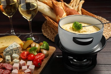 Photo of Fork with piece of broccoli, melted cheese in fondue pot, wine and snacks on wooden table, closeup