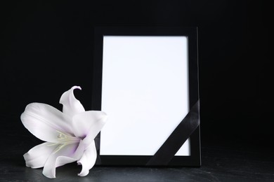 Image of Funeral photo frame with ribbon, white and violet lily on dark table against black background