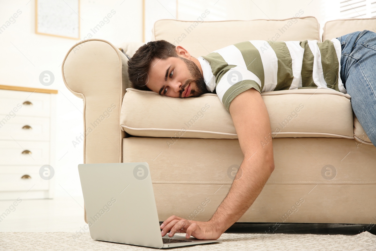 Photo of Lazy young man using laptop while lying on sofa at home