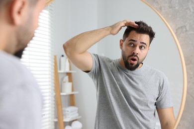 Photo of Emotional man examining his head near mirror in bathroom. Dandruff problem