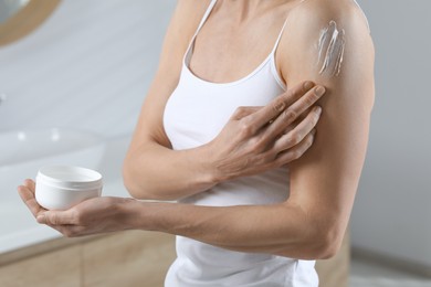 Photo of Woman applying body cream onto arm in bathroom, closeup