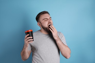 Sleepy young man with cup of coffee yawning on light blue background