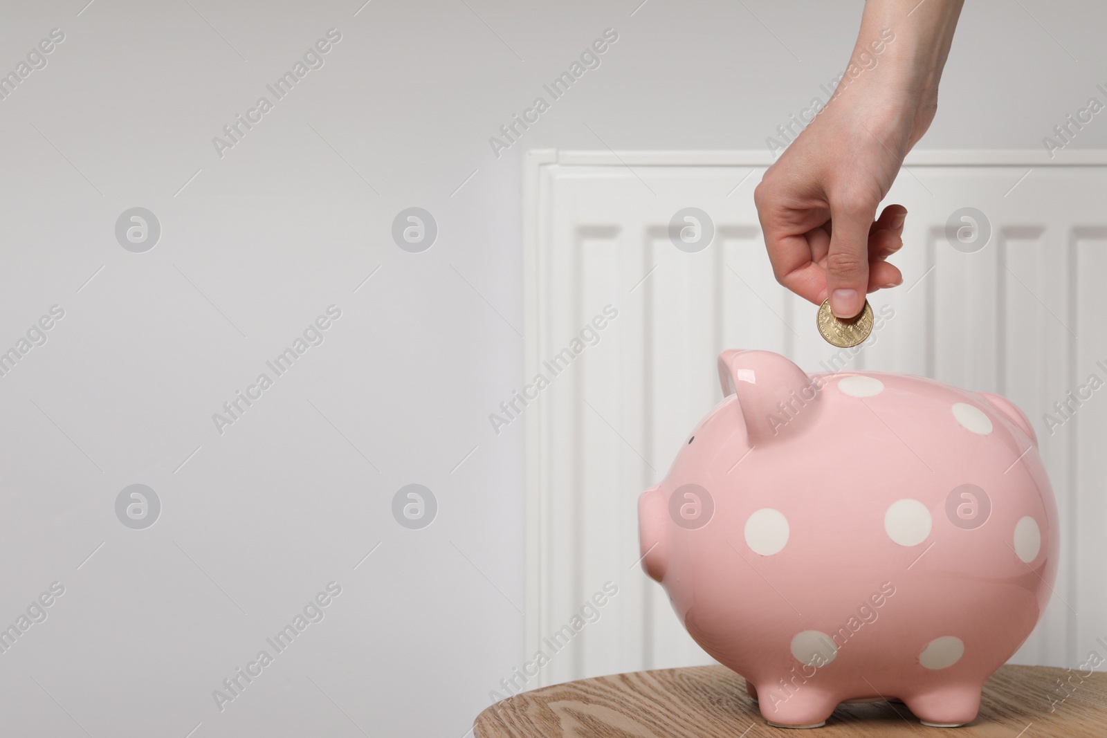 Photo of Woman putting coin into piggy bank near heating radiator, closeup. Space for text