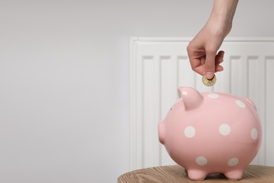 Photo of Woman putting coin into piggy bank near heating radiator, closeup. Space for text