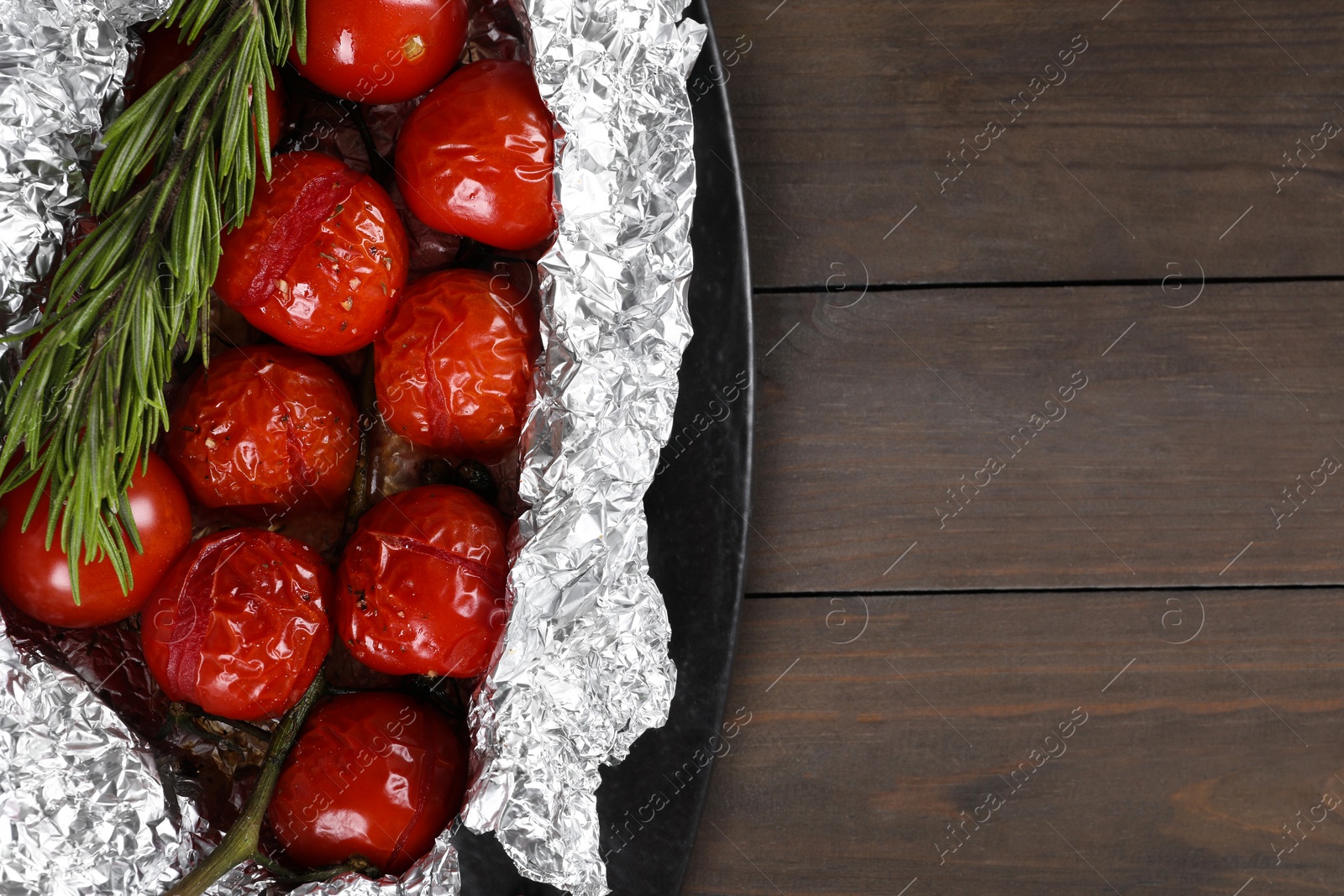 Photo of Aluminum foil with delicious baked tomatoes and rosemary on wooden table, top view. Space for text