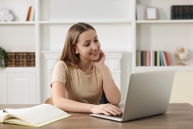 Photo of Online learning. Smiling teenage girl with laptop at wooden table indoors