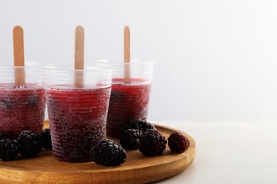 Photo of Tasty blackberry ice pops in plastic cups on white wooden table, space for text. Fruit popsicle