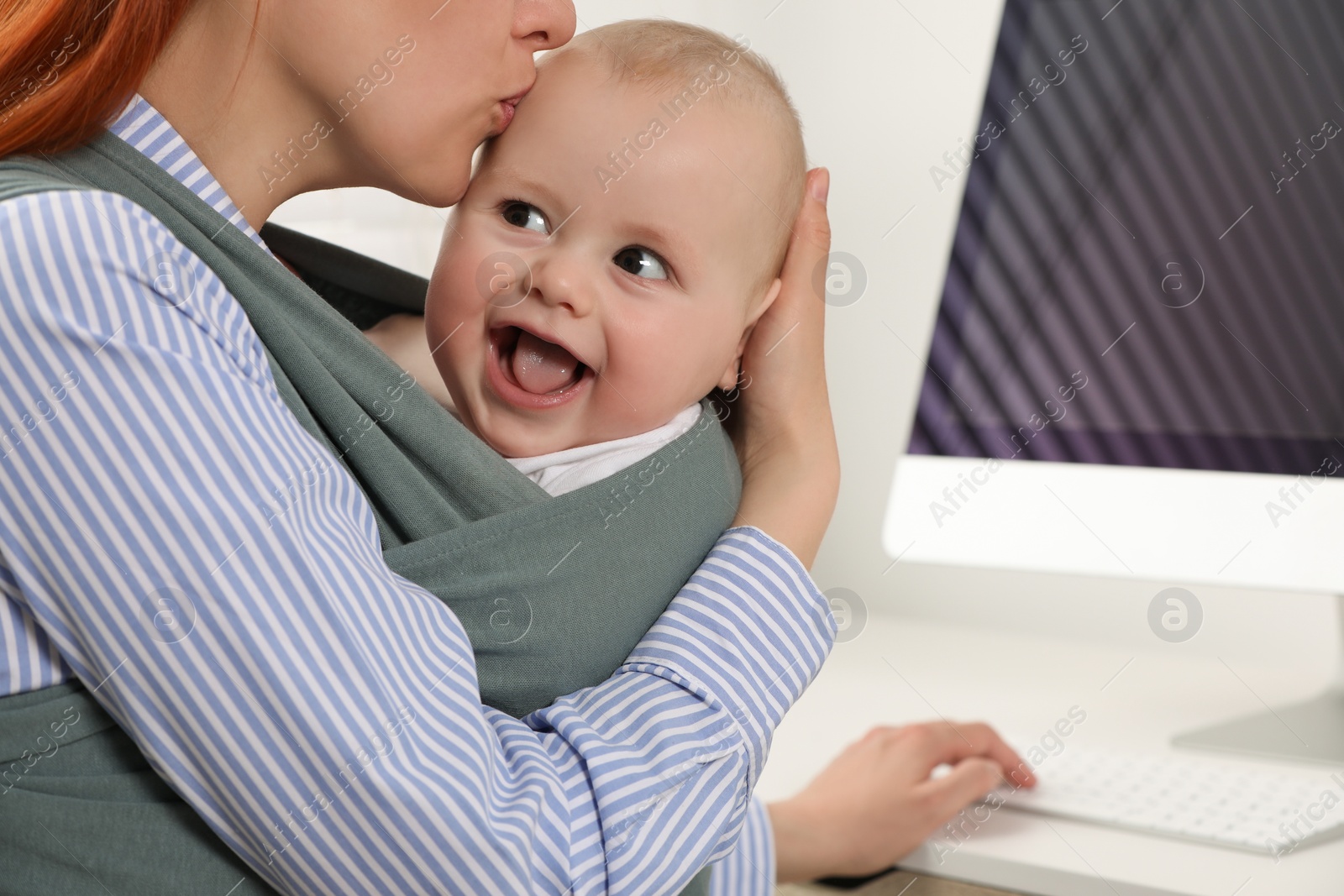 Photo of Mother holding her child in sling (baby carrier) while using computer at workplace