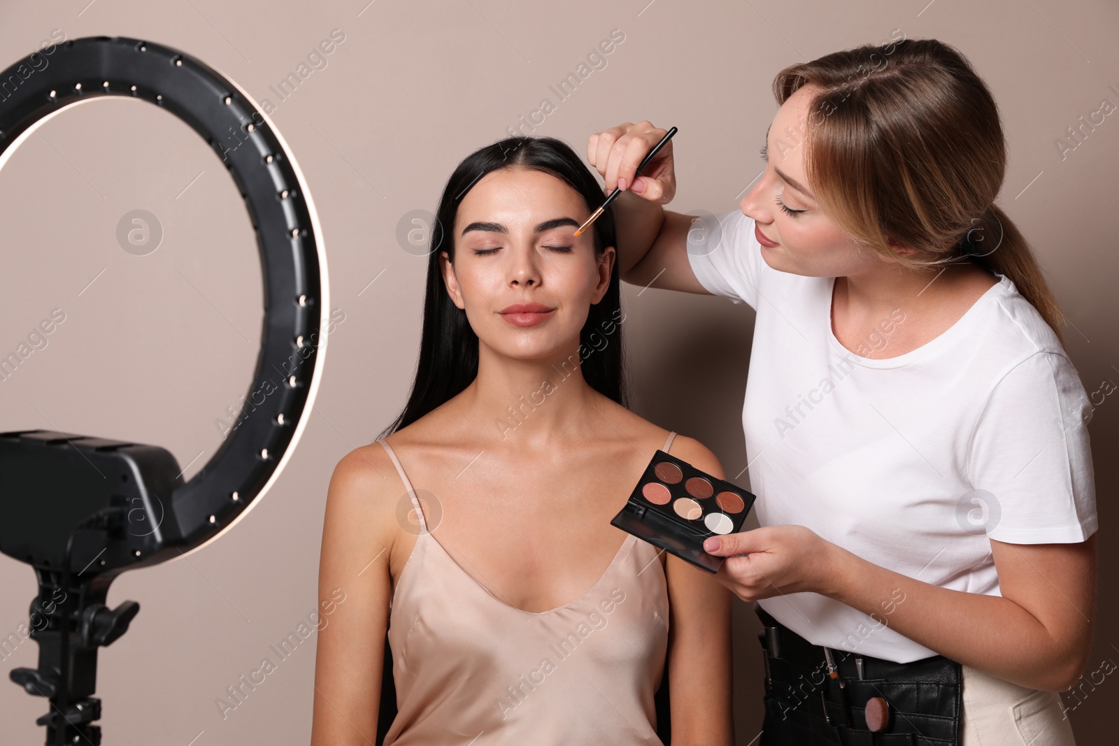 Photo of Professional makeup artist working with beautiful young woman against beige background. Using ring lamp