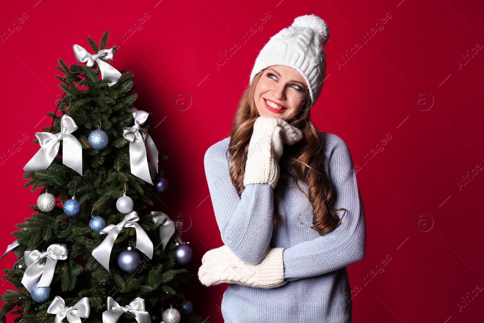 Photo of Beautiful young woman in hat and mittens near Christmas tree on color background