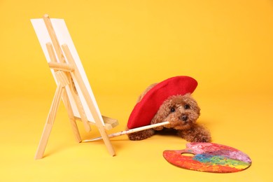 Photo of Cute Maltipoo in red beret holding brush near easel with canvas and palette on orange background. Dog artist