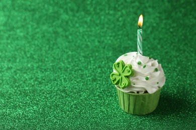 Photo of St. Patrick's day party. Tasty cupcake with clover leaf topper and burning candle on shiny green background. Space for text