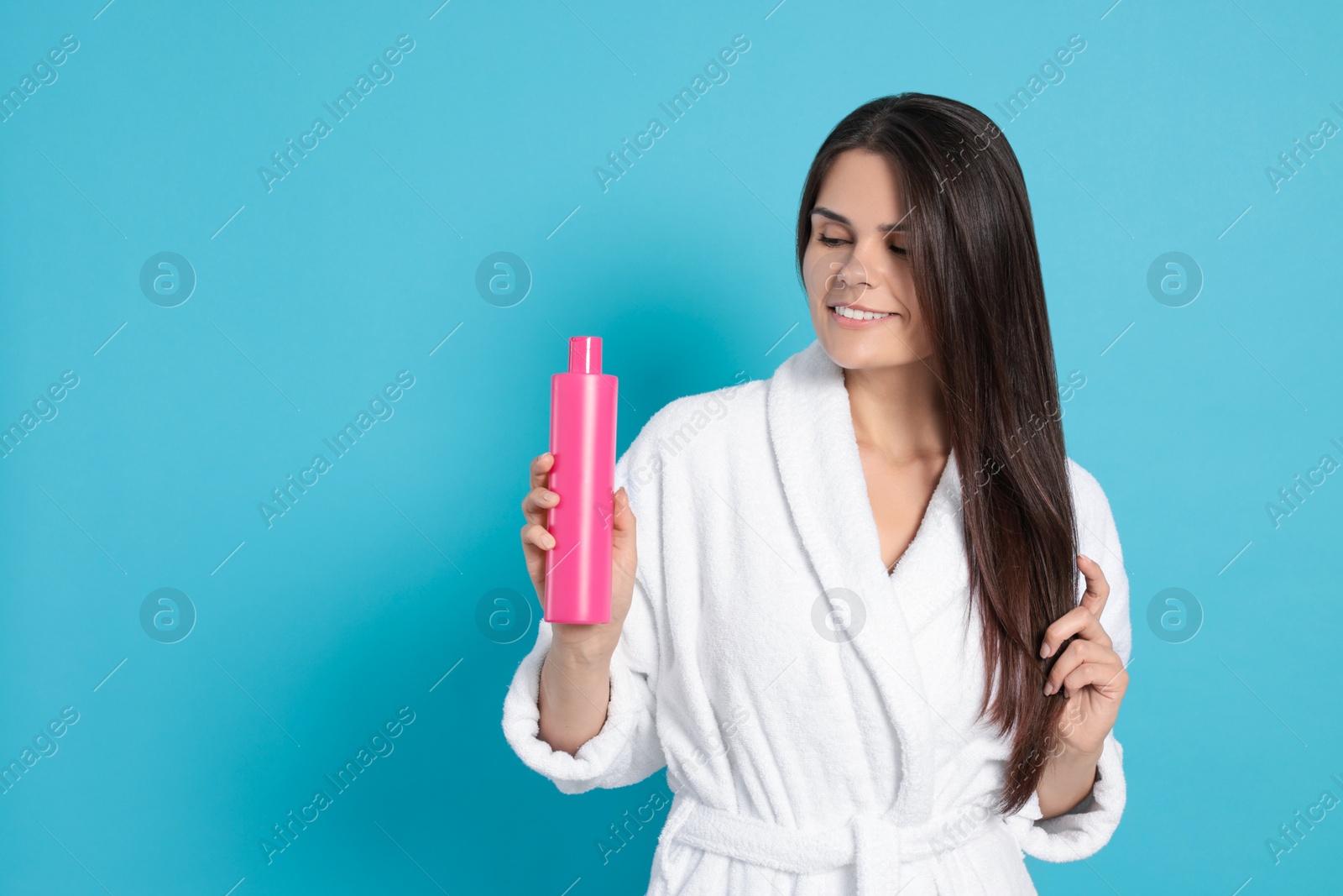 Photo of Beautiful young woman in bathrobe holding bottle of shampoo on light blue background, space for text