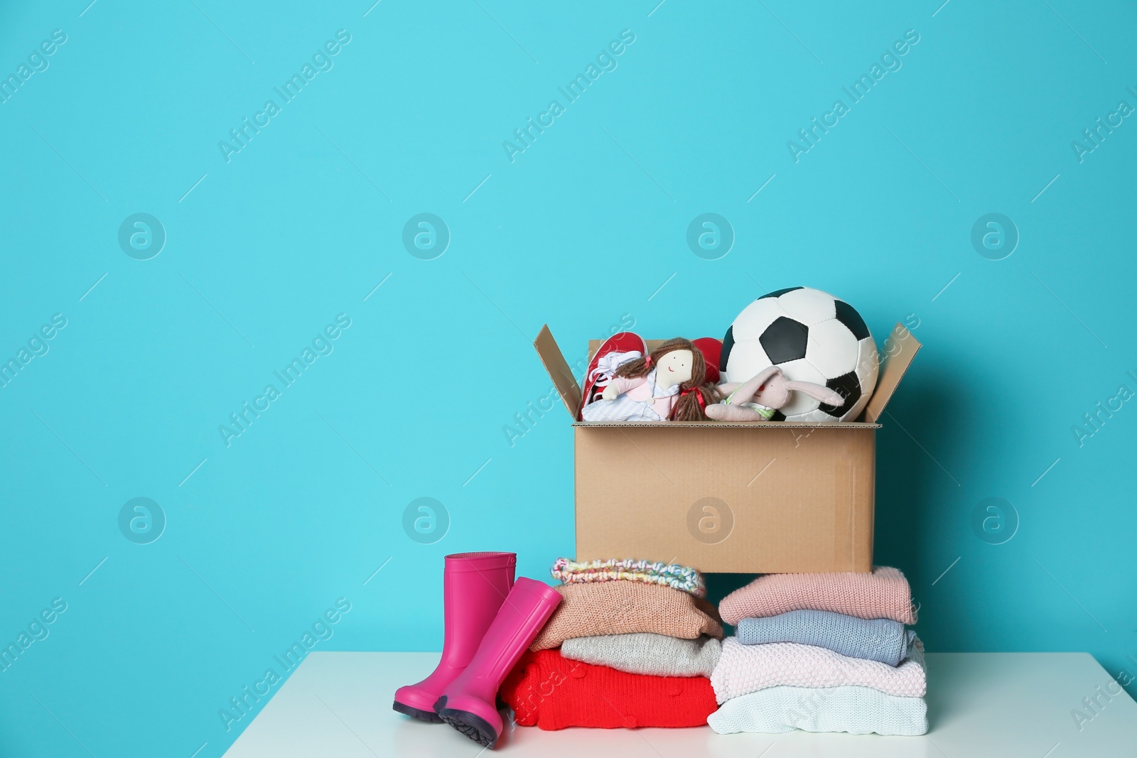 Photo of Donation box with toys, knitted clothes and rubber boots on table against color background. Space for text