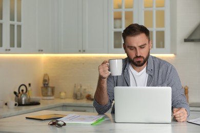 Photo of Young man with cup of drink working on laptop at home, space for text
