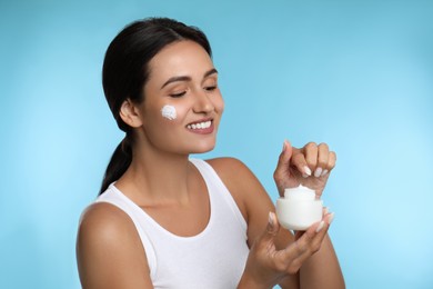 Young woman applying facial cream on light blue background