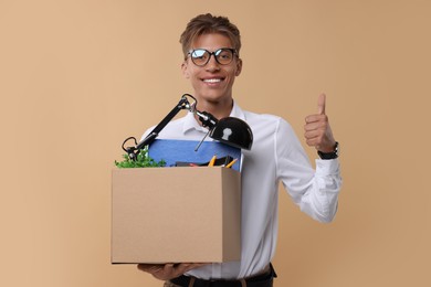 Photo of Happy unemployed young man with box of personal office belongings showing thumb up on beige background