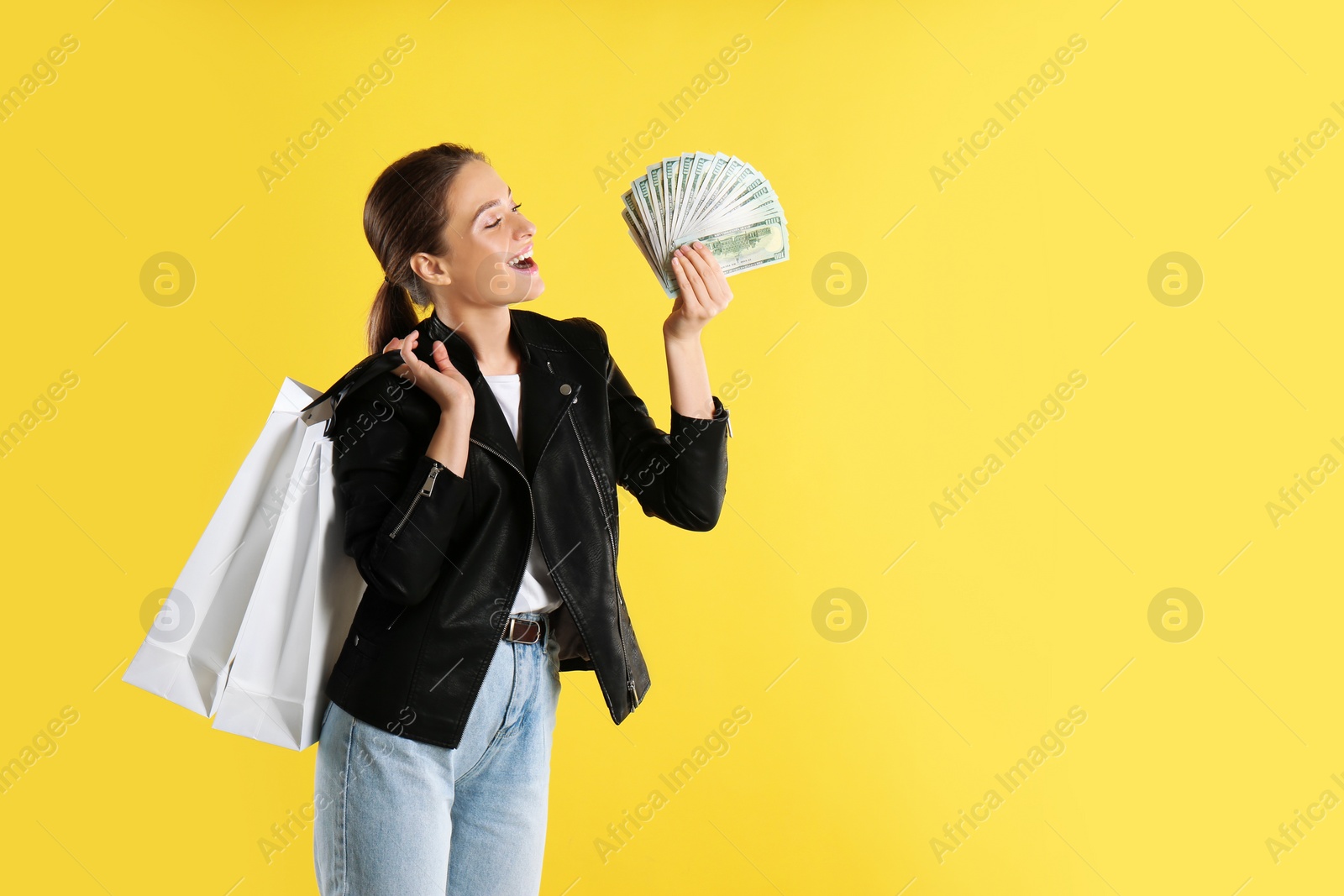 Photo of Emotional young woman with money and shopping bags on yellow background. Space for text