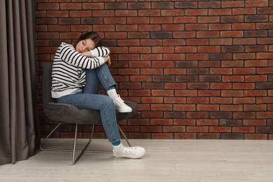 Photo of Sad young woman sitting on chair near brick wall indoors, space for text