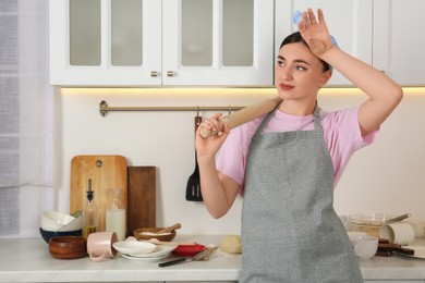 Photo of Tired housewife with rolling pin in kitchen. Dirty dishware, food and utensils on messy countertop behind her