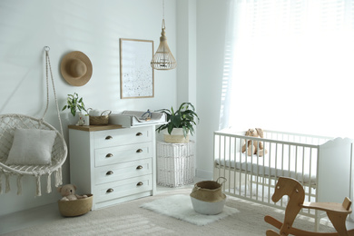 Photo of Chest of drawers with changing tray and pad near comfortable cradle in baby room. Interior design