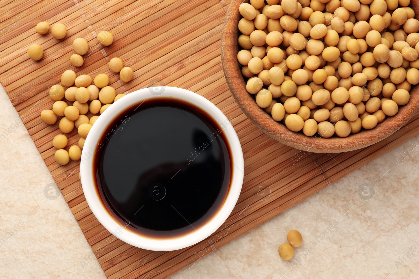 Photo of Soy sauce in bowl and soybeans on beige table, flat lay