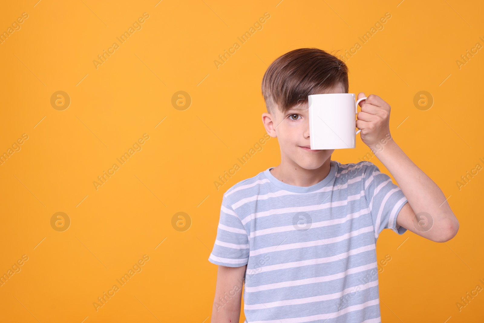 Photo of Cute boy covering eye with white ceramic mug on orange background, space for text