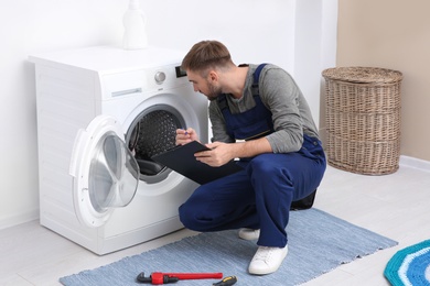 Photo of Young plumber with clipboard near washing machine in bathroom