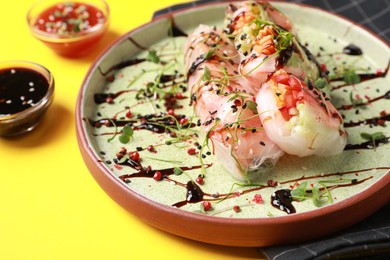 Plate of delicious spring rolls with sauce and microgreens on yellow background, closeup