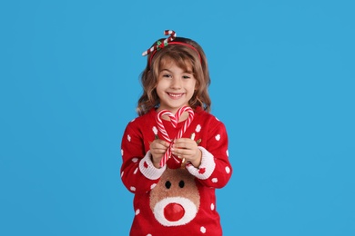 Photo of Cute little girl in Christmas sweater making heart shape with candy canes against blue background