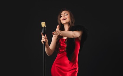 Beautiful young woman in stylish dress with microphone singing on black background