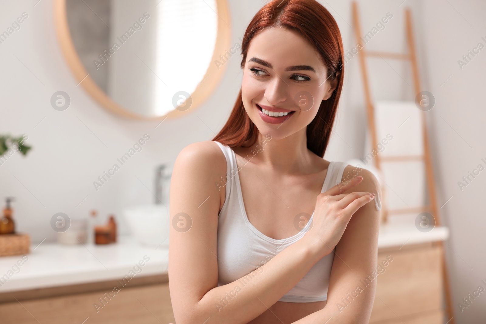 Photo of Beautiful young woman applying body cream onto shoulder in bathroom, space for text