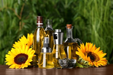 Photo of Many different bottles with cooking oil, sunflower seeds and flowers on wooden table against blurred green background