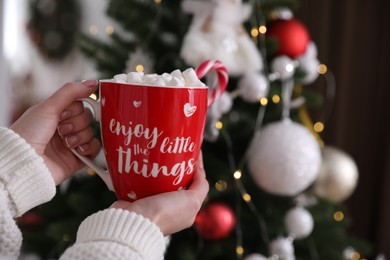 Woman with cup of delicious hot drink near Christmas tree indoors, closeup