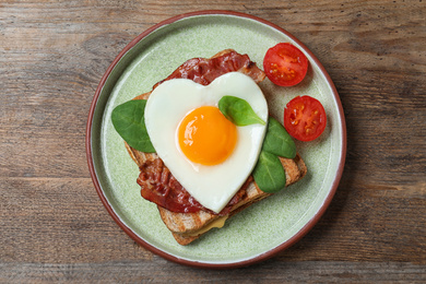 Tasty sandwich with heart shaped fried egg and  bacon on wooden table, top view