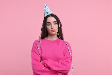 Sad woman in party hat with blower and streamers on pink background