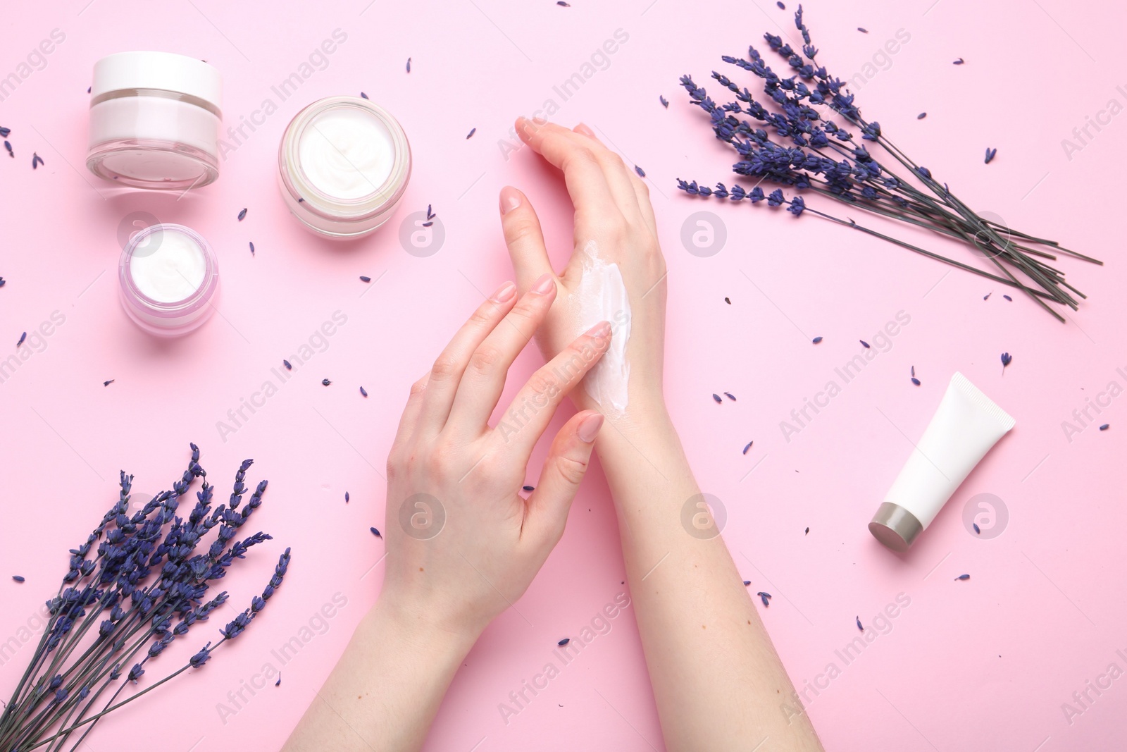 Photo of Woman applying hand cream and lavender flowers on pink background, top view