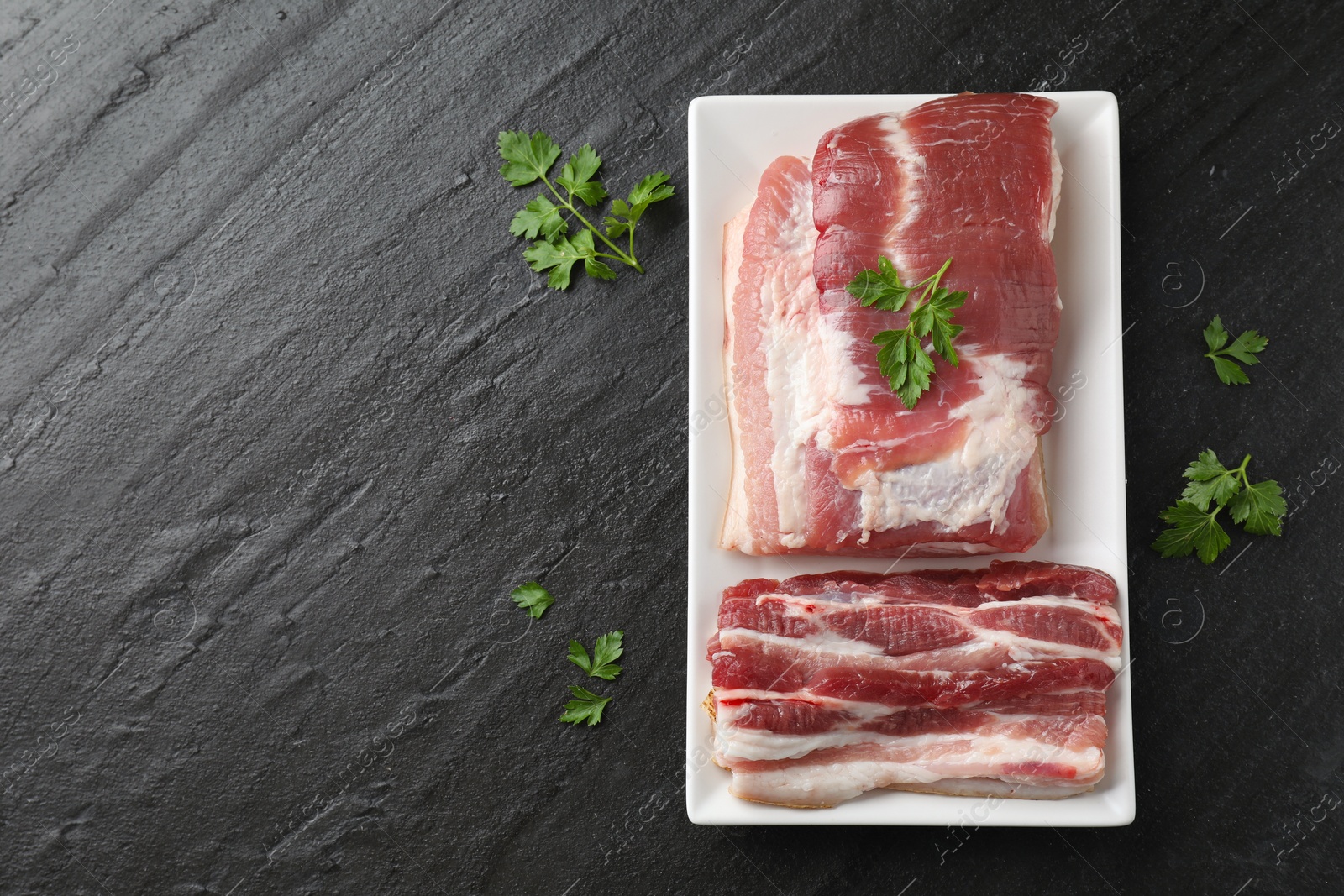 Photo of Pieces of raw pork belly and parsley on black textured table, top view. Space for text