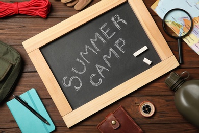 Chalkboard with text SUMMER CAMP and camping equipment on wooden background, flat lay