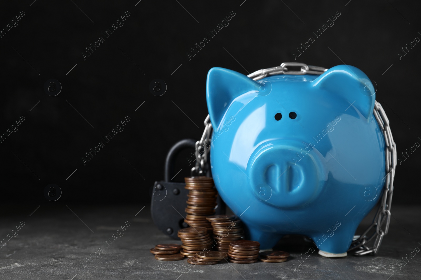 Photo of Piggy bank with steel chain, padlock and coins on dark grey stone table against black background, space for text. Money safety concept